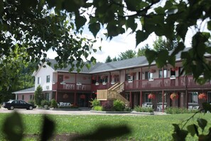 A two-story building with a red exterior and a balcony. There is a car parked in front and a grassy area in the foreground. - Motel Le Radisson de Val-David (Val David)