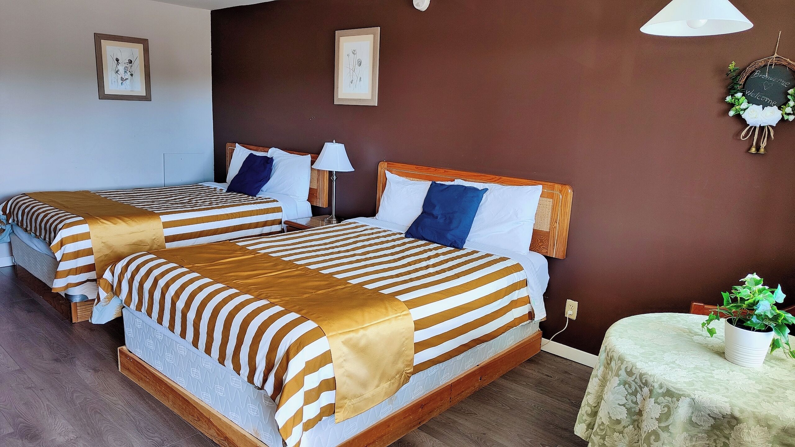A hotel room with two beds, each with striped bedding and pillows, a bedside table with a lamp, and a small table with a potted plant.