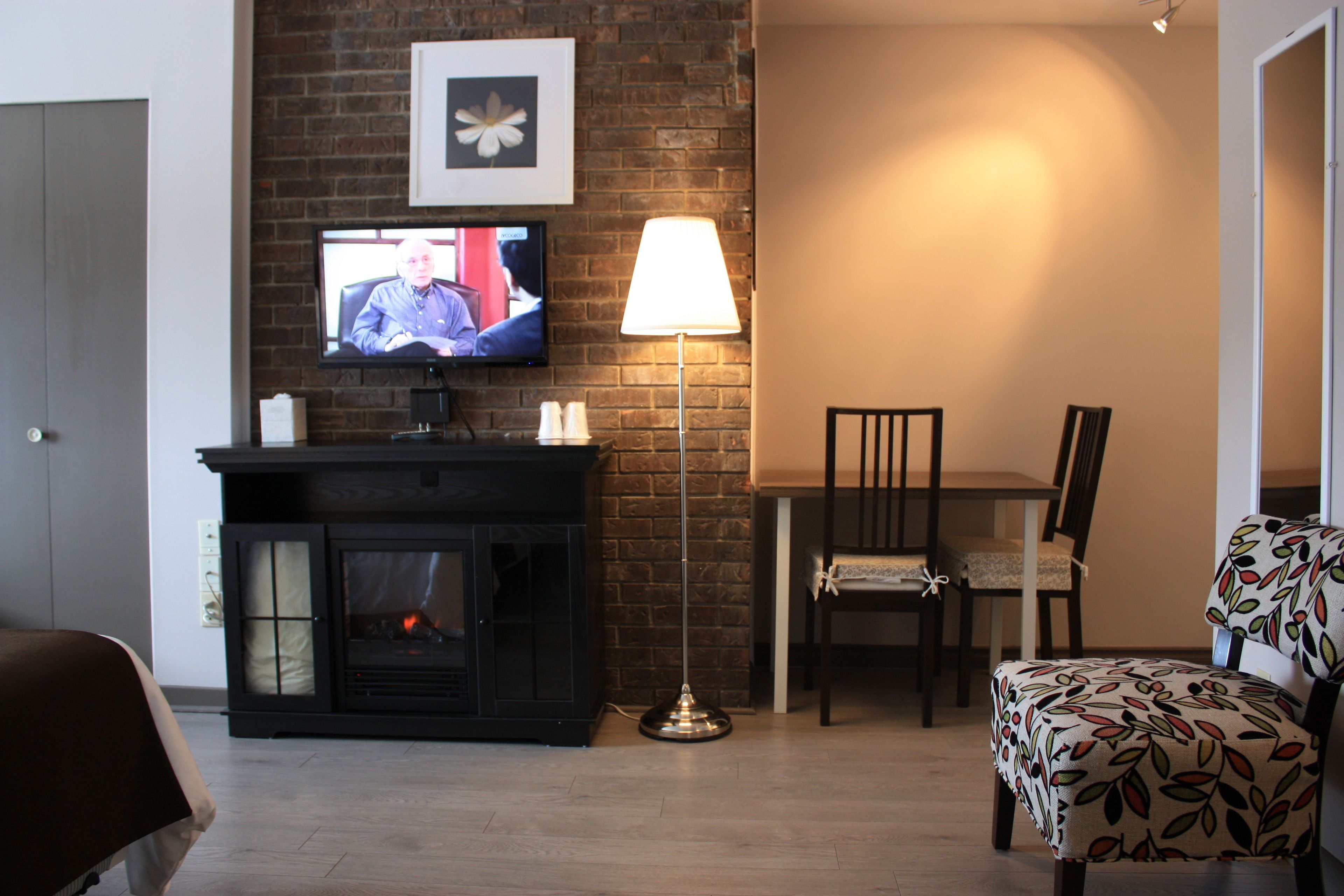 A living room with a fireplace, a television mounted on a brick wall, a table with chairs, and a patterned armchair.