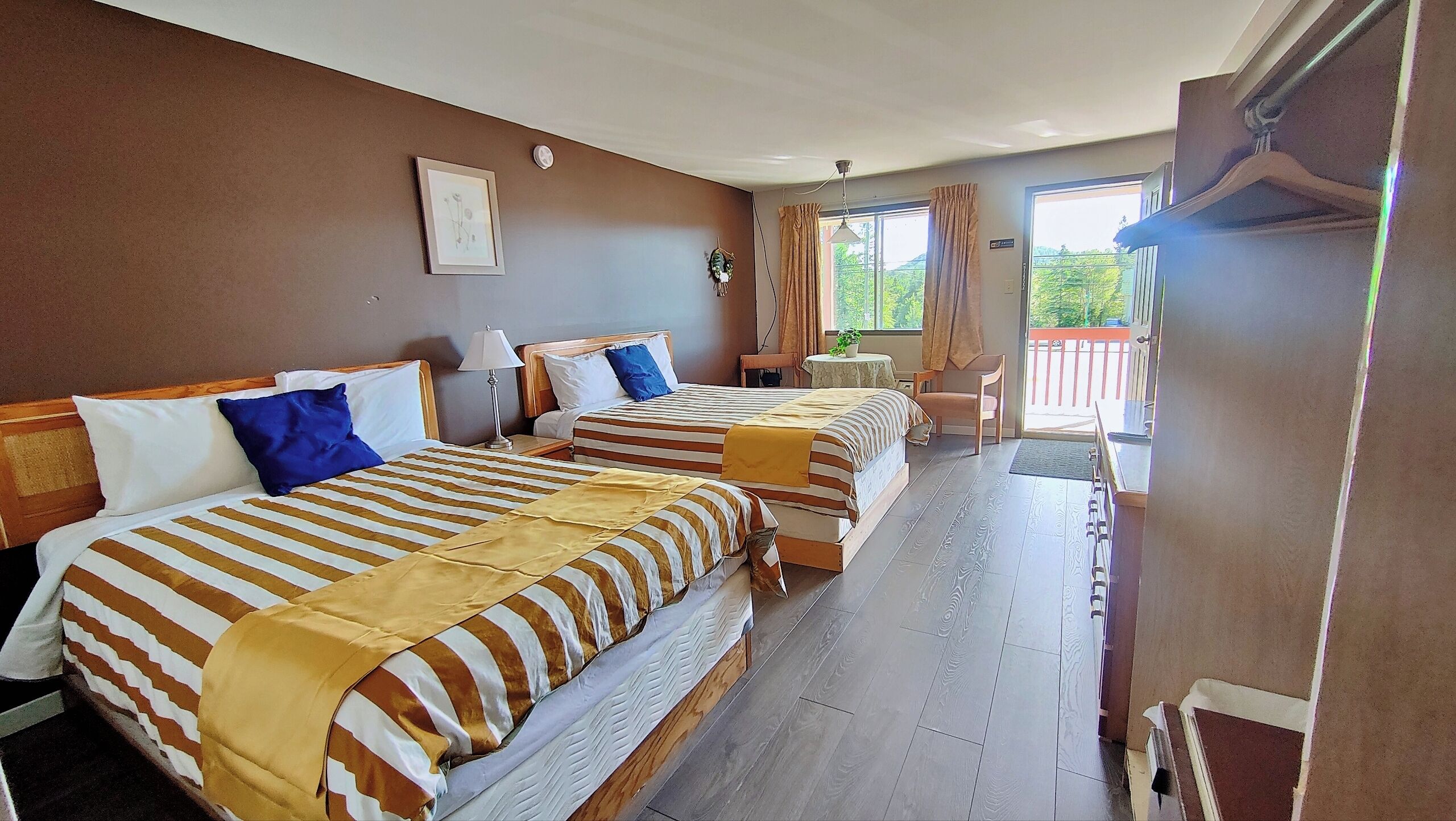 A hotel room with two beds, a small table, a chair, a window with curtains, and a view of greenery outside.