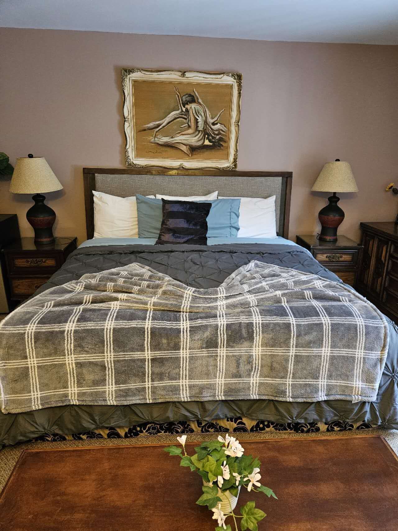 Una cama bien hecha con un edredón escocés, dos lámparas de noche, una obra enmarcada sobre la cama y una planta pequeña en el suelo.
