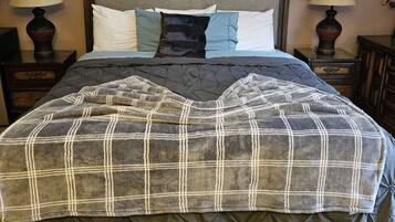 Una cama bien hecha con un edredón escocés, dos lámparas de noche, una obra enmarcada sobre la cama y una planta pequeña en el suelo.