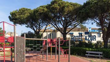 A playground with climbing structures and a bench, with a hotel named "HOTEL AZUR" in the background.