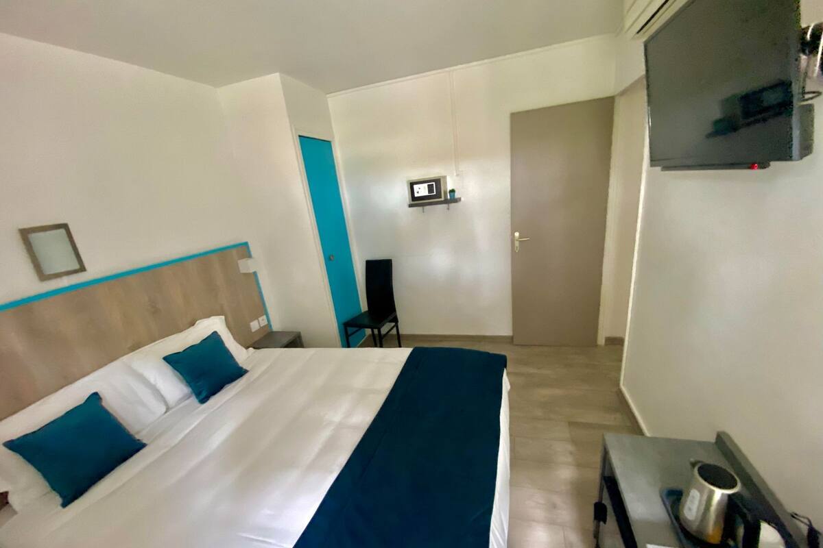 A hotel room with a bed, a desk with a kettle, a TV, and a door.