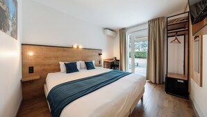 Une chambre d’hôtel moderne dotée d’un grand lit, d’une tête de lit en bois, d’un bureau avec une chaise et offrant une vue sur l’extérieur grâce à une porte vitrée.