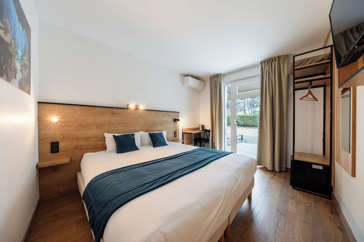 A modern hotel room with a large bed, a wooden headboard, a desk with a chair, and a view through a glass door to the outdoors.
