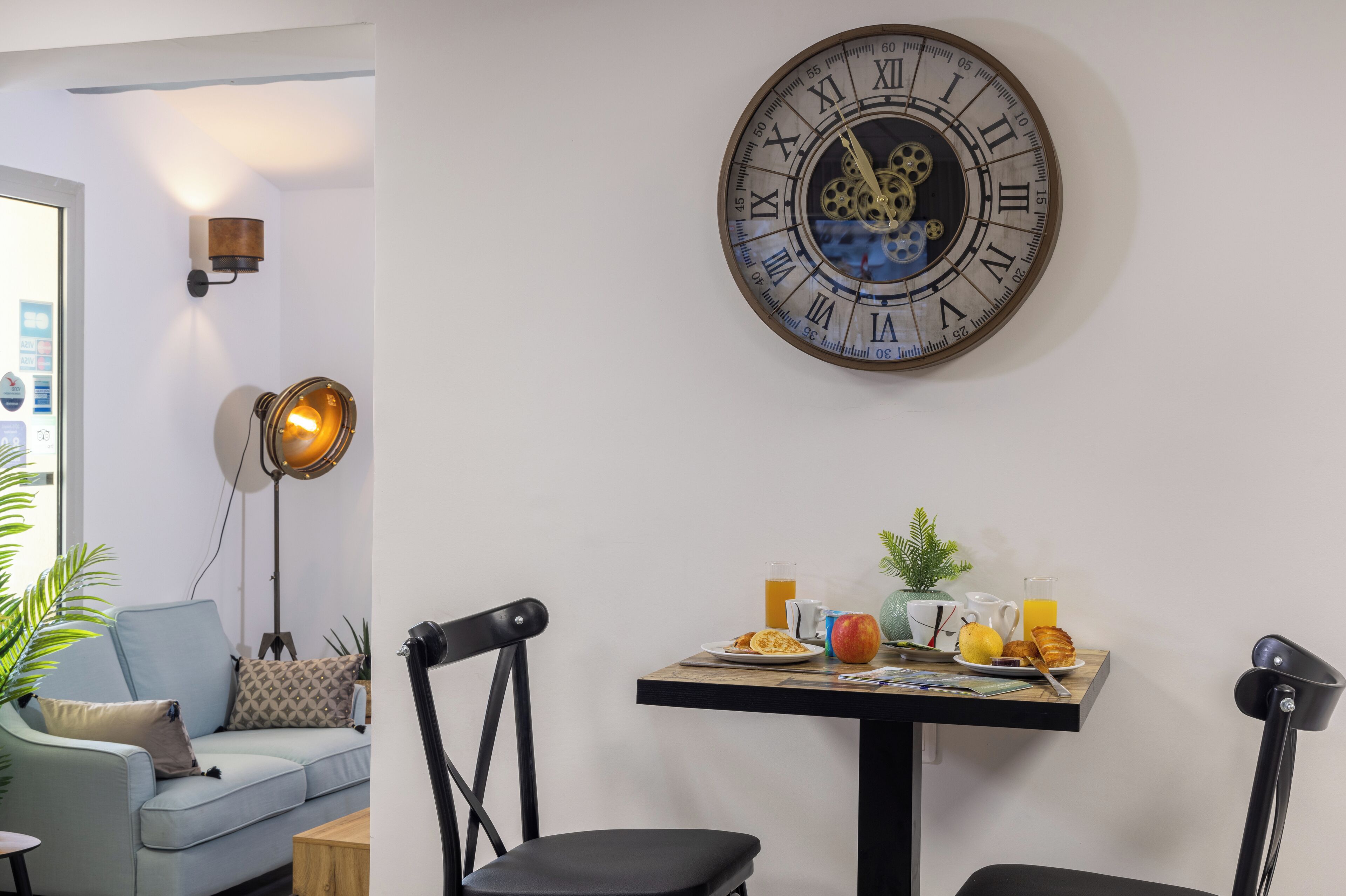 A room with a round wall clock, a table with breakfast items, a sofa, and a lamp.