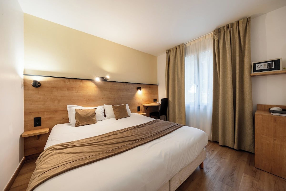 A hotel room with a large bed, wooden headboard, bedside tables with lamps, a desk, and a view of the outdoors through a window with curtains.