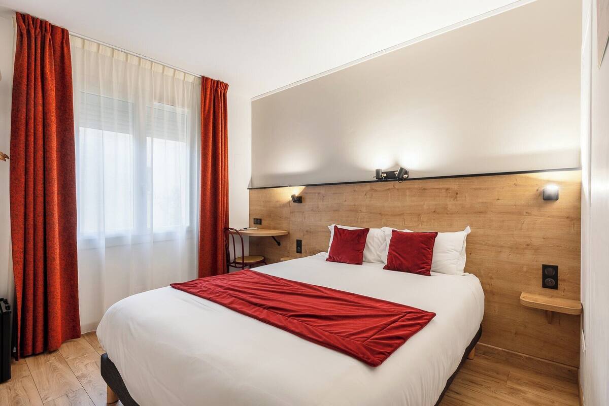 A hotel room with a large bed, red and white bedding, wooden headboard, bedside tables, and a window with sheer curtains.