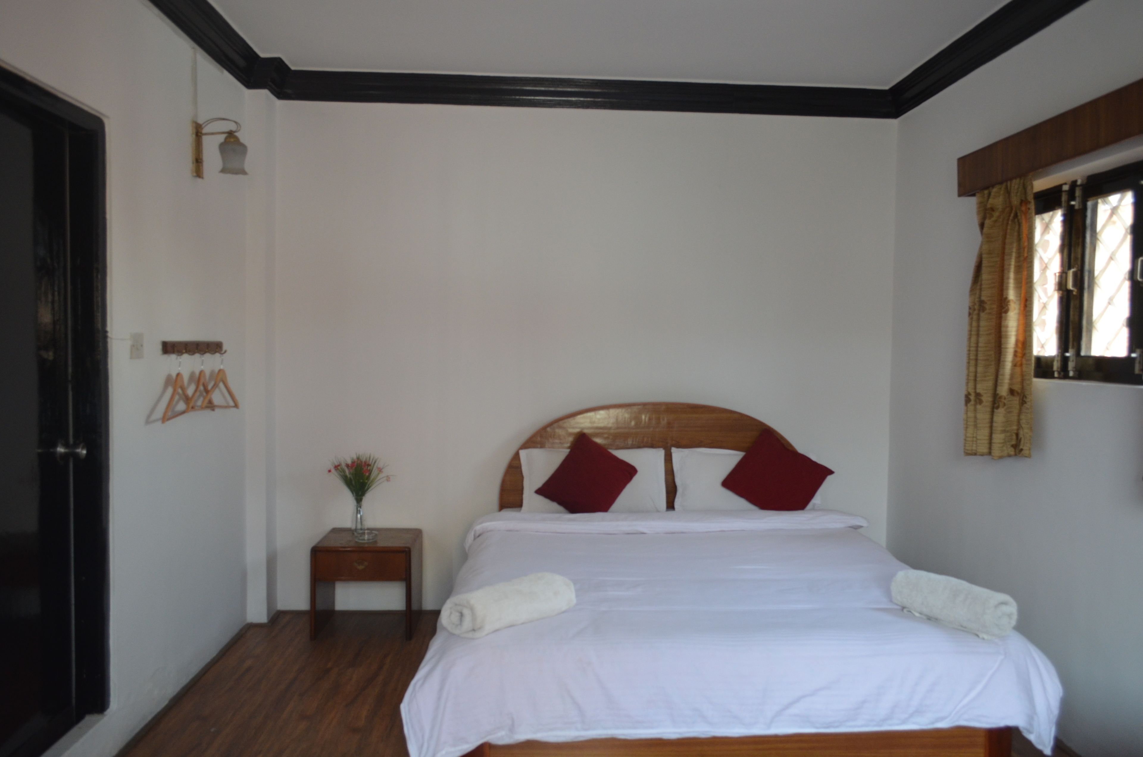 A neatly made bed with white linens and two red pillows, a small bedside table with a vase of flowers, and a wooden headboard.