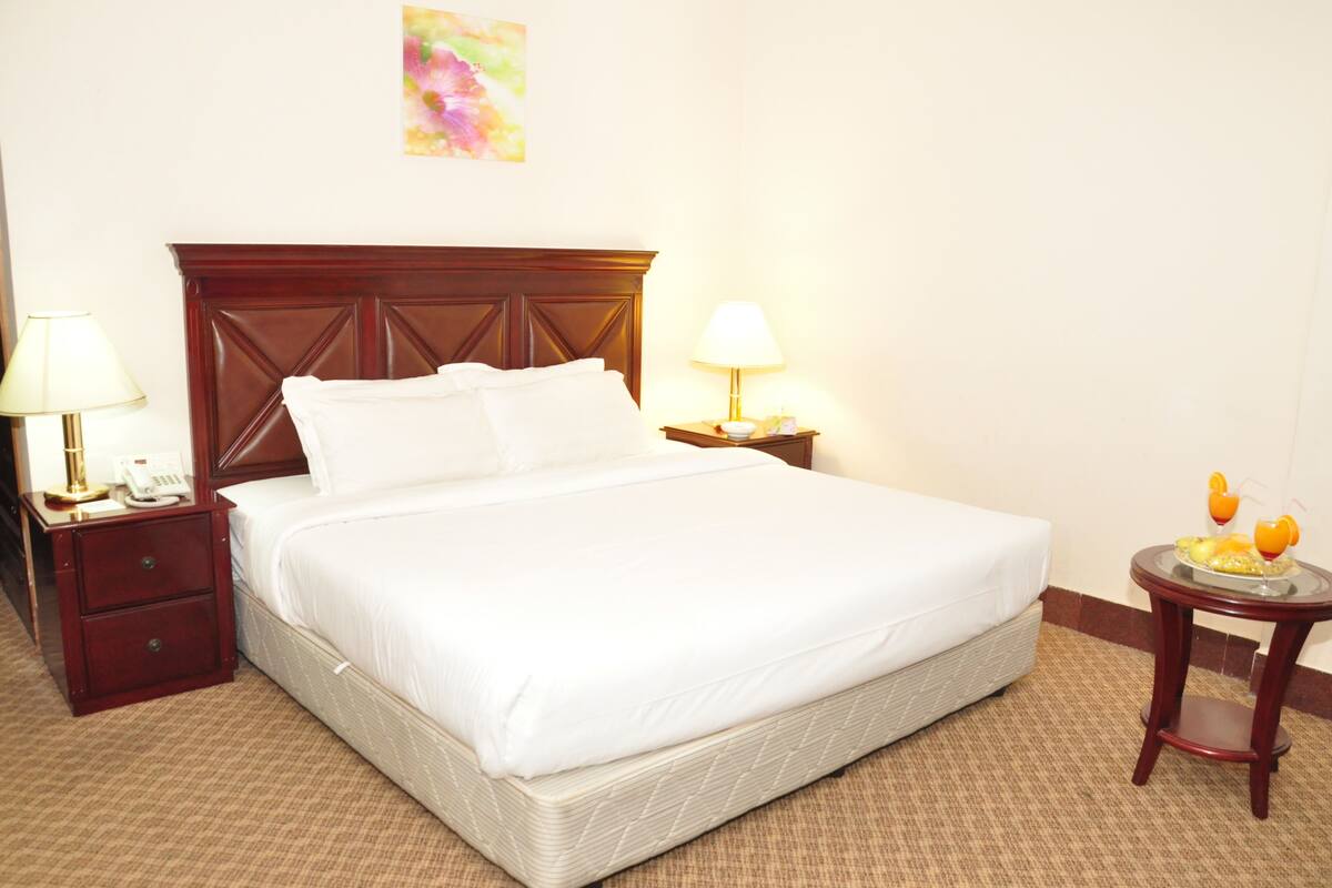 A neatly made bed with white linens, a wooden headboard, two bedside tables with lamps, and a small round table with fruit.