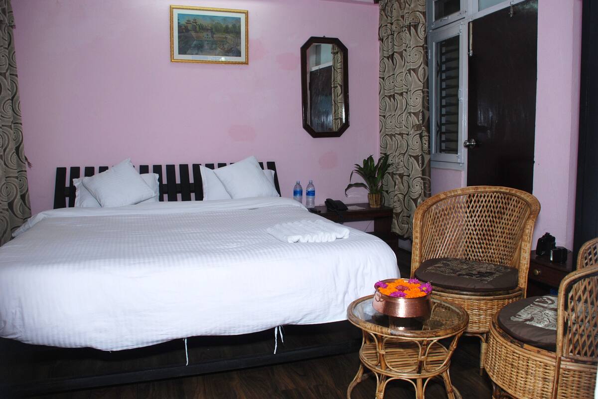 A bedroom with a bed, two wicker chairs, a small table with a flower vase, a mirror, and a door.