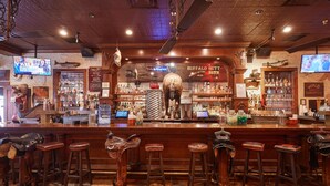 A bar with wooden stools, a counter displaying various bottles, and a television mounted on the wall. - Stockyards Hotel (Fort Worth)