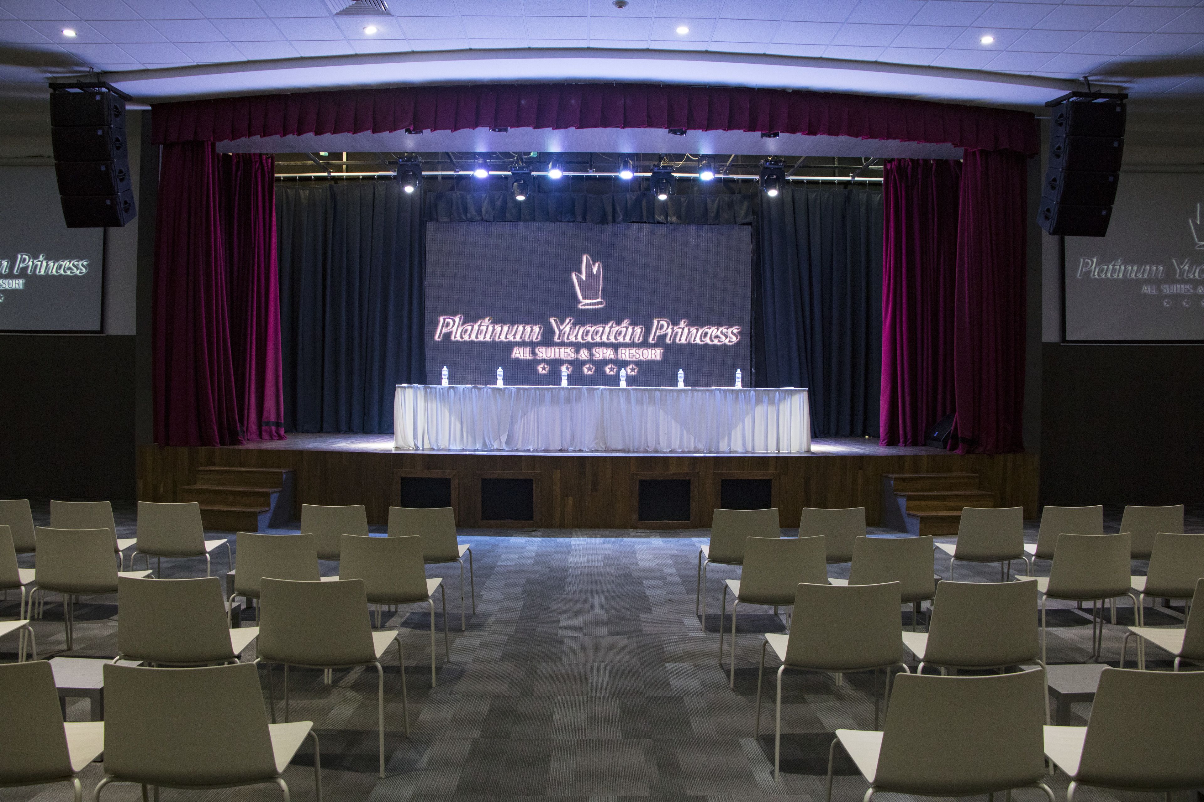 Uma sala de conferências com palco, cadeiras e uma tela exibindo "Platinum Yucatán Princess All Suites & Spa Resort".