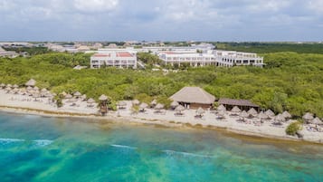 Vista aérea de un resort con playa, construcciones de techo de paja y un edificio grande con múltiples balcones.