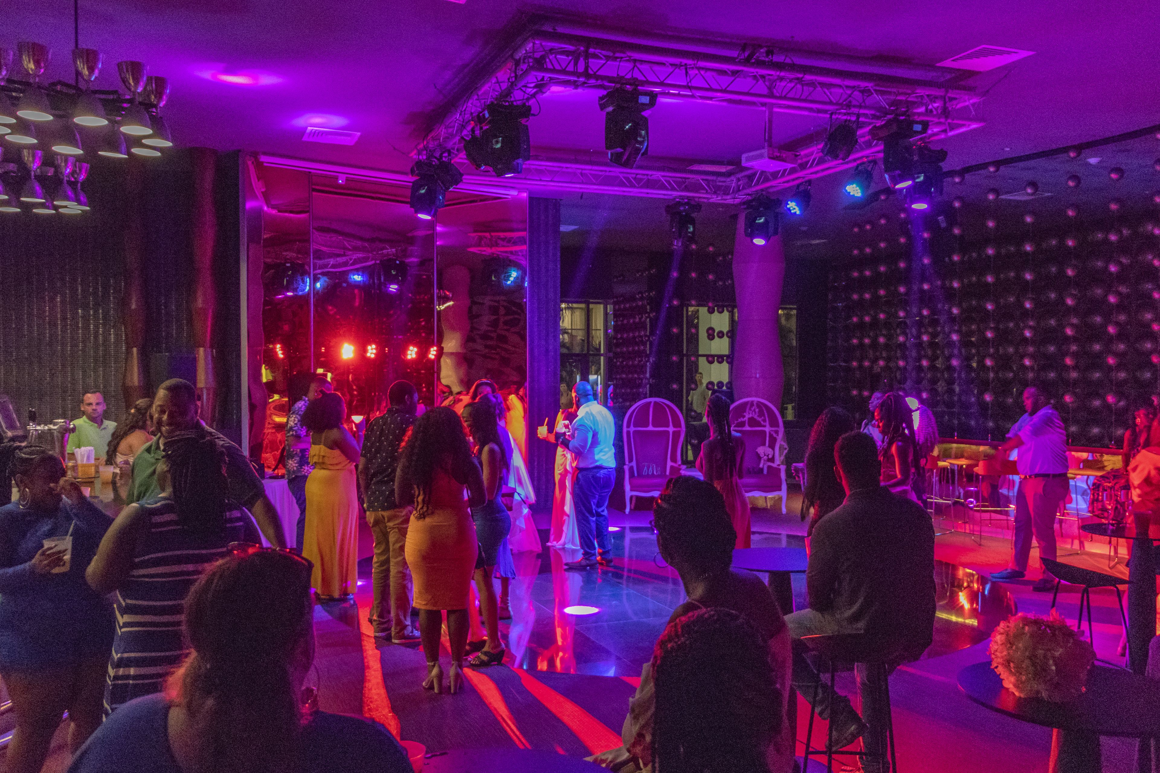 A crowded indoor event with people standing and sitting, a stage with performers, and a wall of spherical decorations.