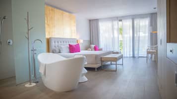 A modern hotel room with a large bed, a separate seating area, a bathtub, and a view of greenery outside.