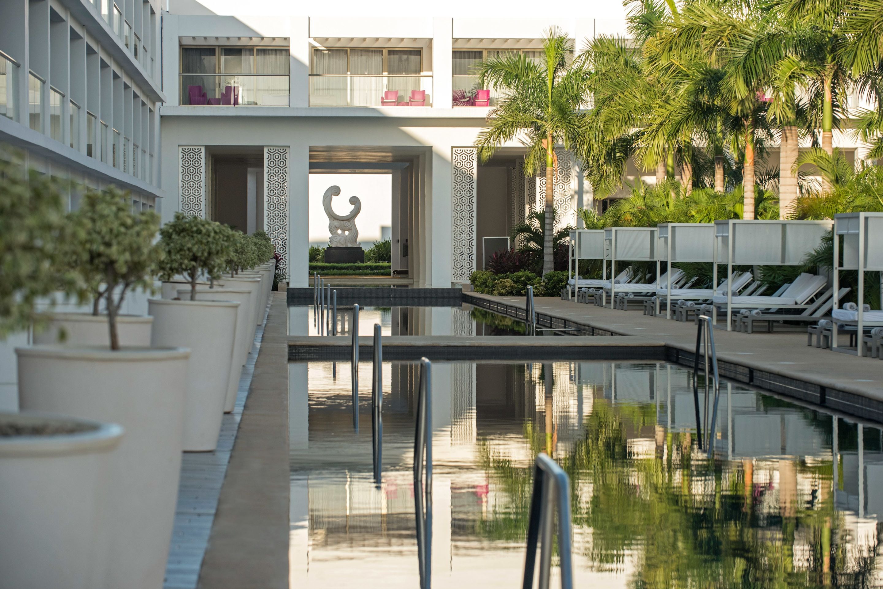 Uma área de piscina moderna em hotel, com espreguiçadeiras, palmeiras e uma escultura central.