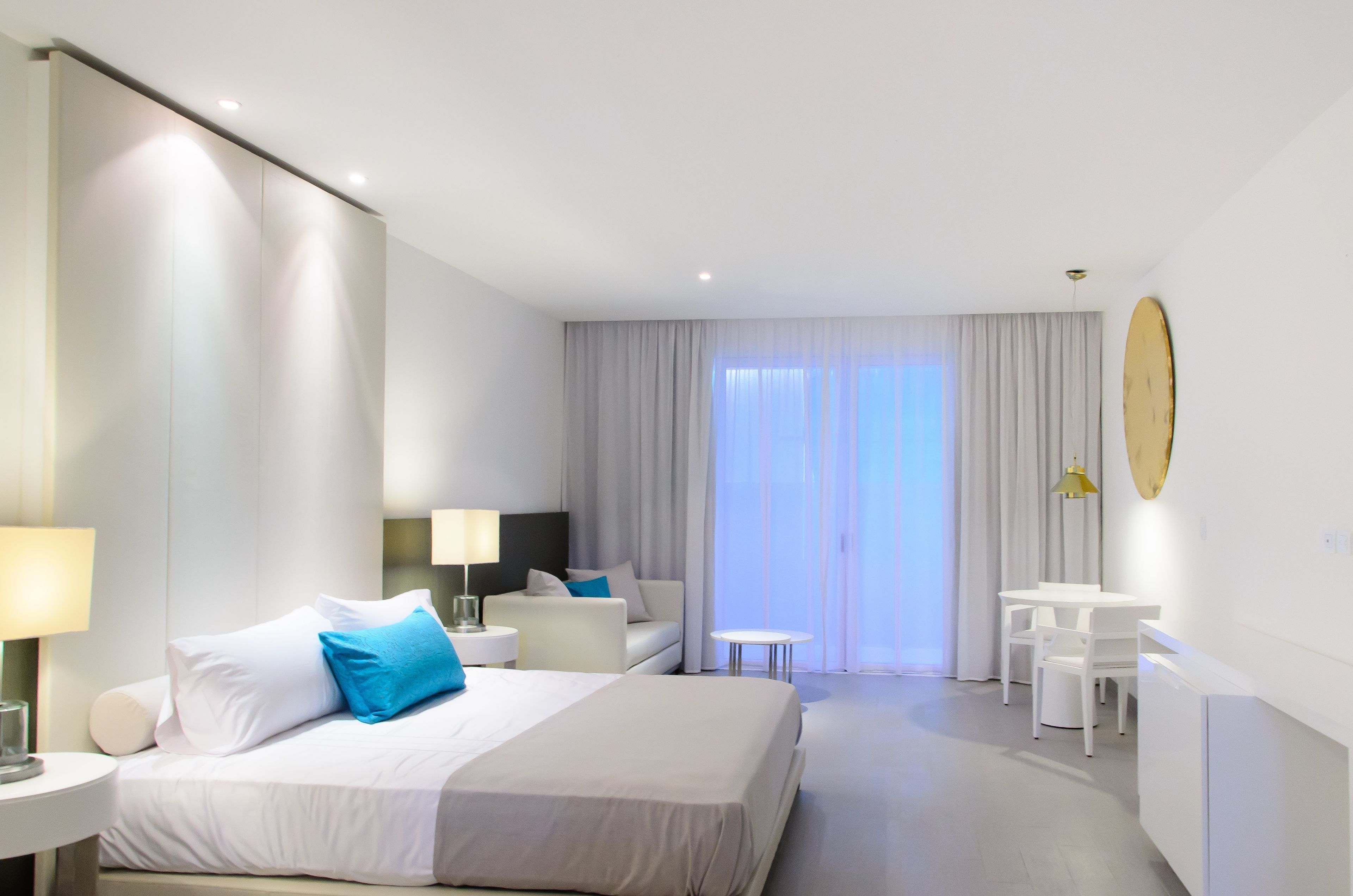 A modern hotel room with a large bed, a desk, and a chair. There is a window with curtains and a view of the outside.
