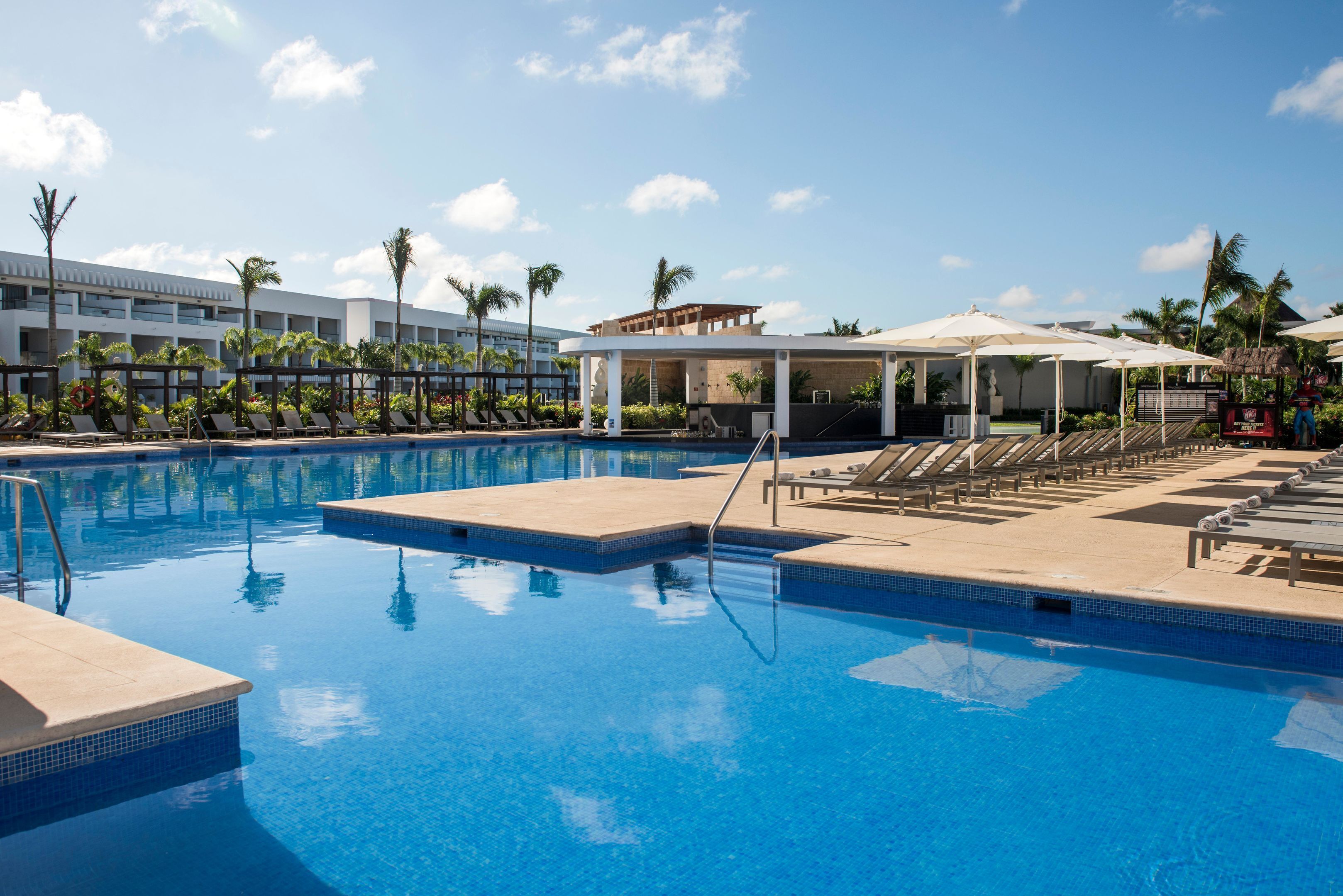 A swimming pool with lounge chairs and umbrellas, surrounded by palm trees and a building with multiple floors.