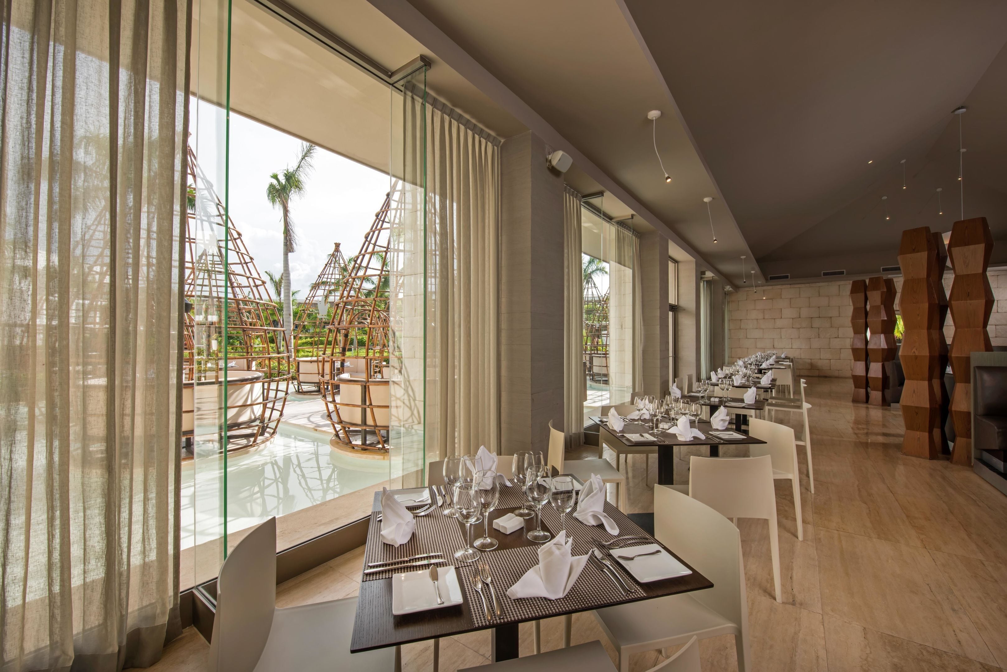 A modern dining area with tables set for service, large windows with curtains, and a view of outdoor structures.