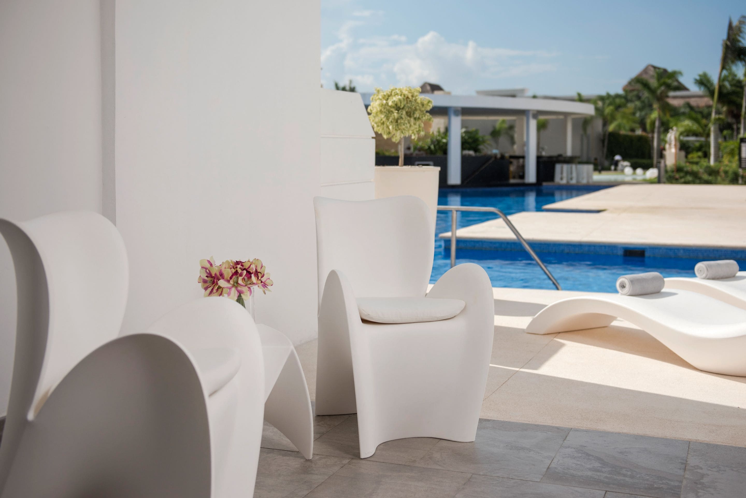 Un área moderna junto a la piscina con sillones blancos y una pequeña mesa con flores.