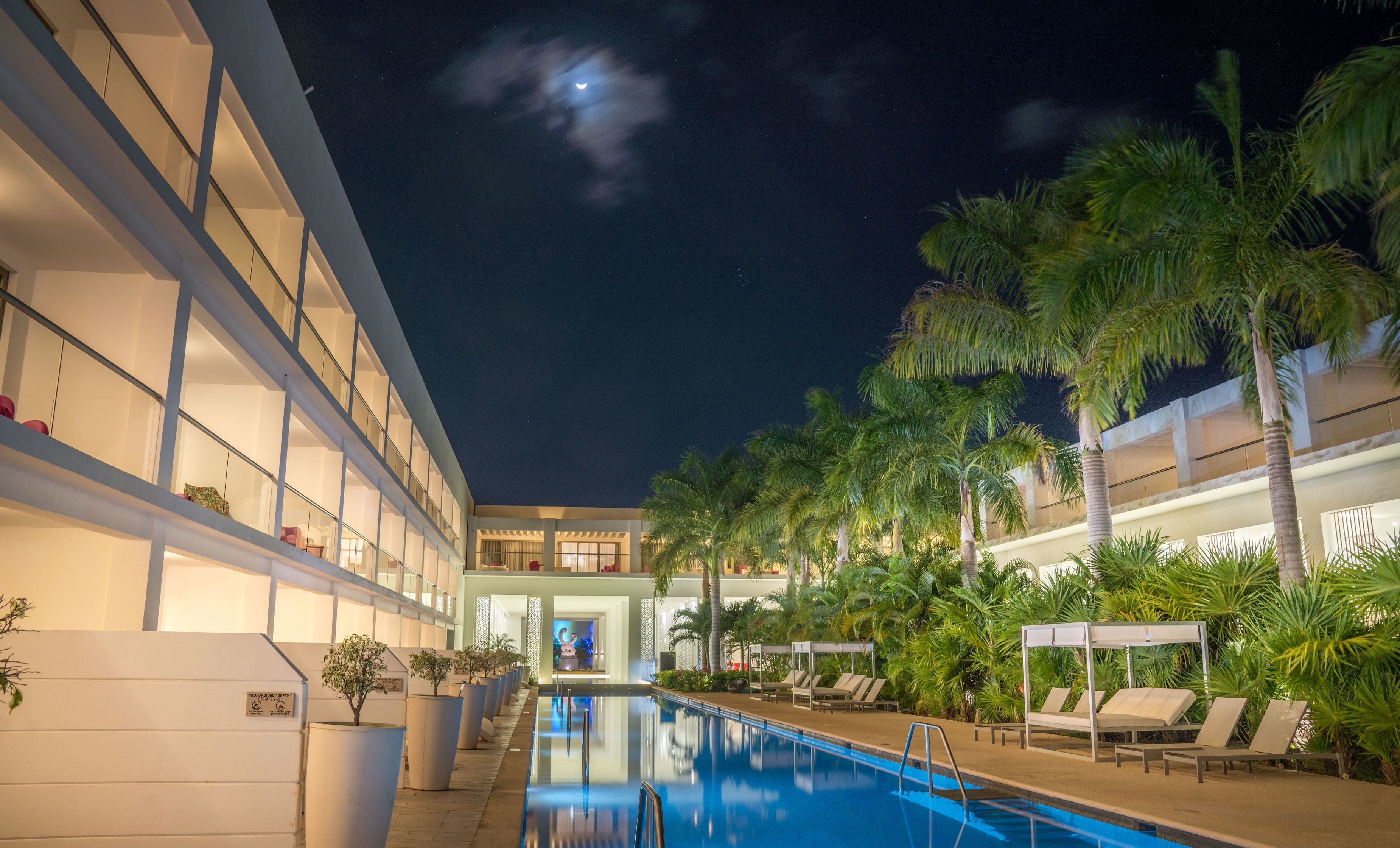 A swimming pool area with lounge chairs and palm trees, surrounded by a multi-story building with balconies.