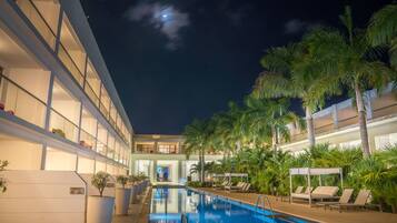 A swimming pool area with lounge chairs and palm trees, surrounded by a multi-story building with balconies.