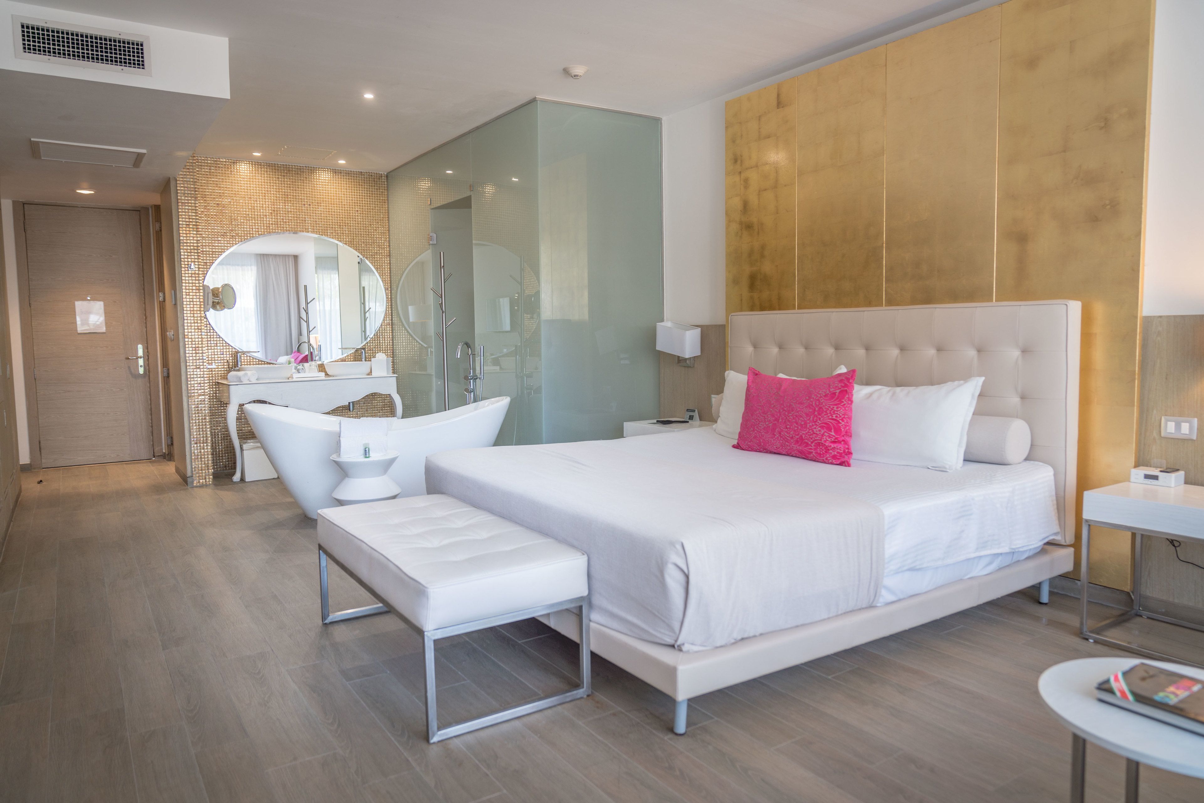 A modern hotel room with a large bed, a sitting area, a vanity with a mirror, and a shower area.