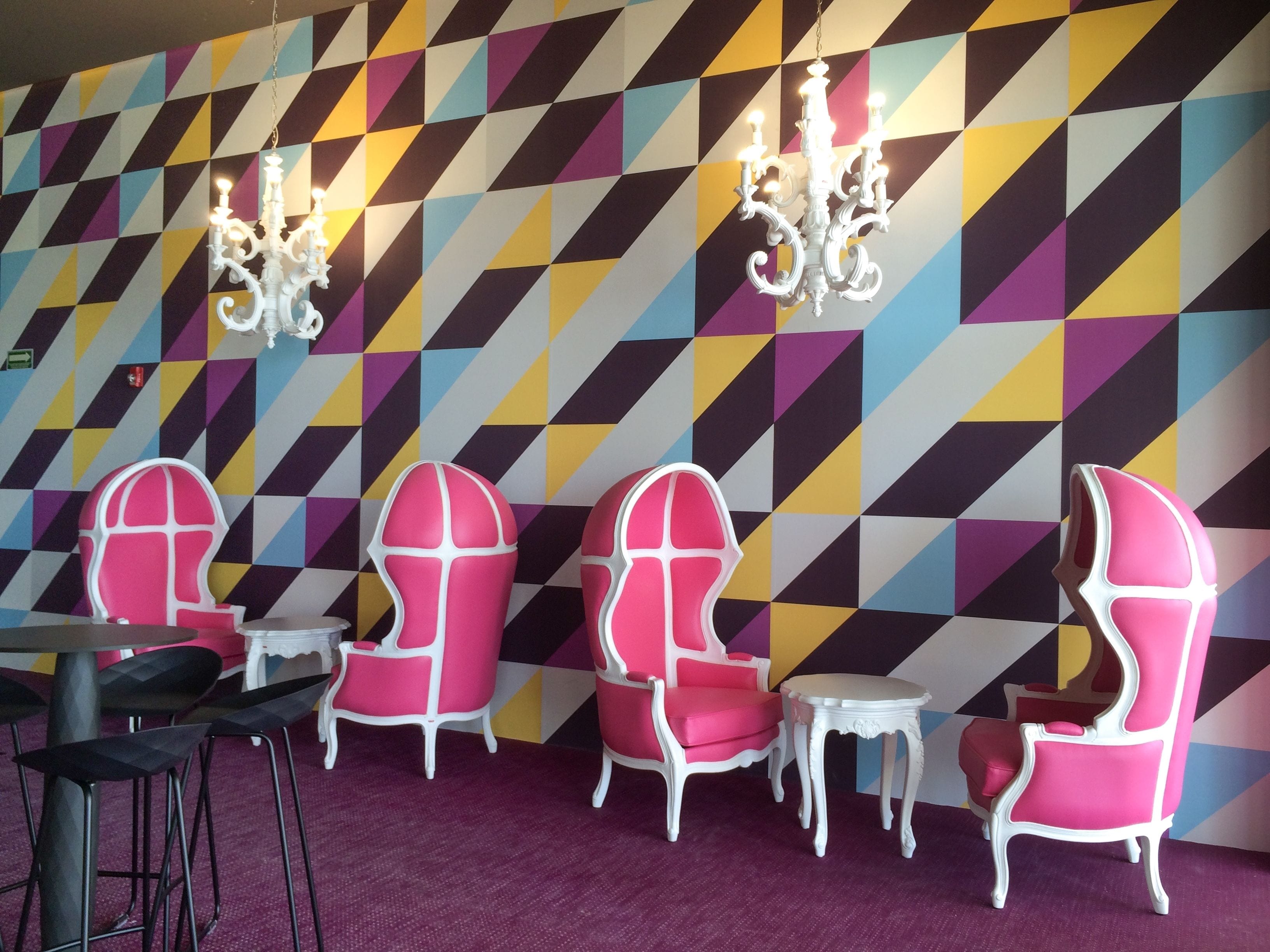 A room with a geometric patterned wall, white ornate chandeliers, and pink and white chairs with small round tables.
