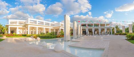 Un hotel moderno con piscina, mobiliario exterior y un área cubierta con sombrillas.