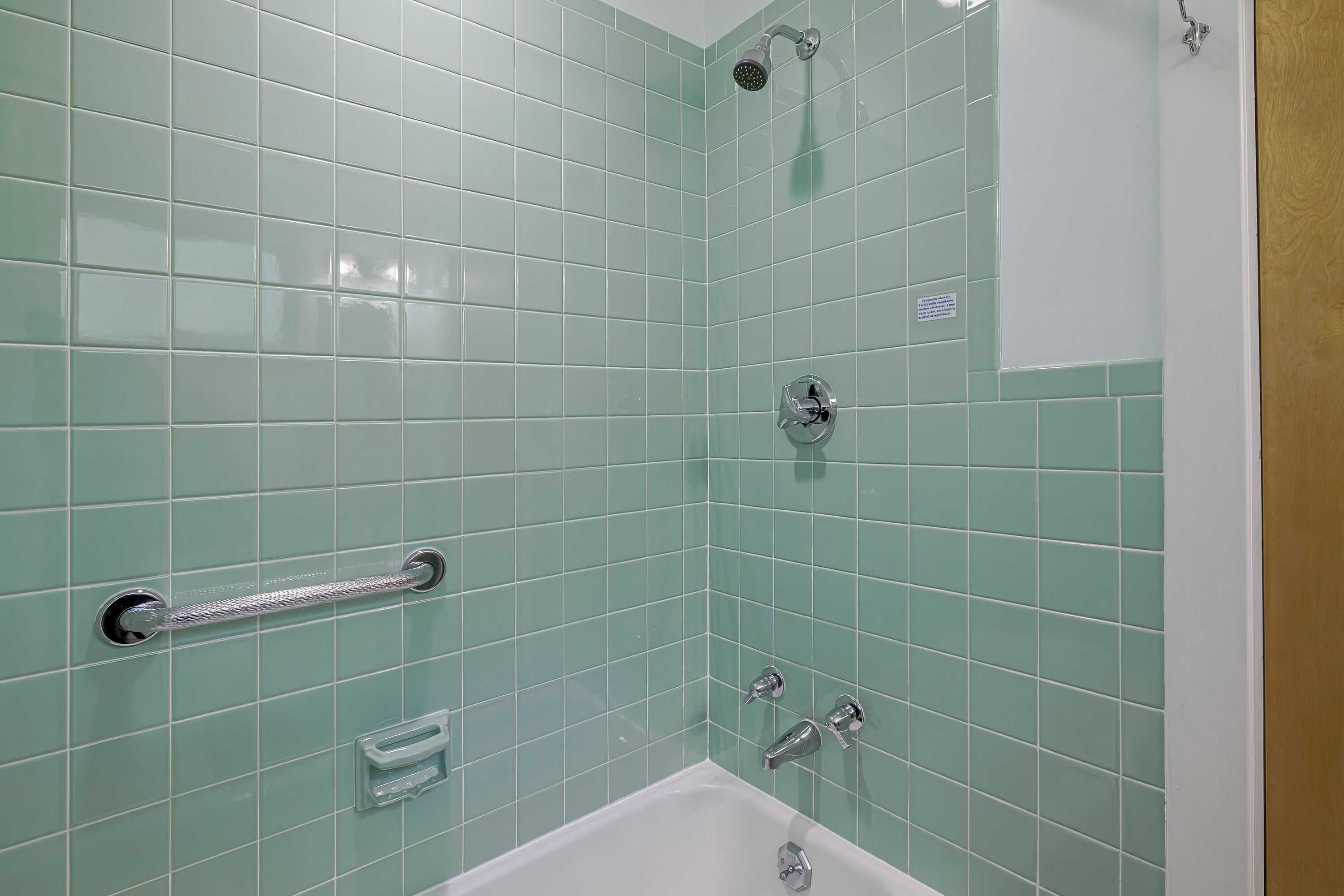 Un baño con azulejos verdes, una bañera blanca, un cabezal de ducha y un asidero.