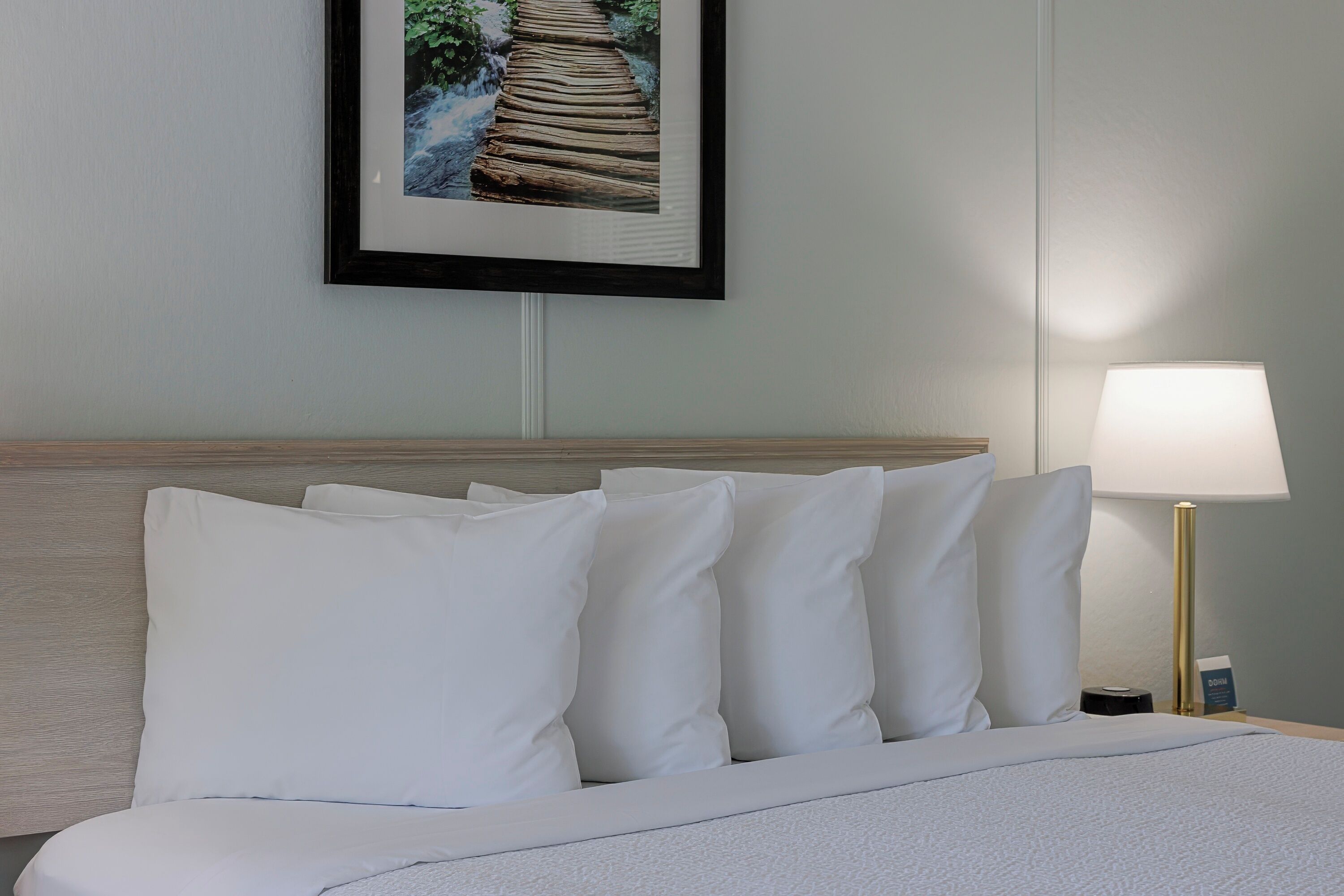 A neatly made bed with white bedding and pillows, a framed picture of a wooden bridge over a stream, and a bedside lamp with a gold base.