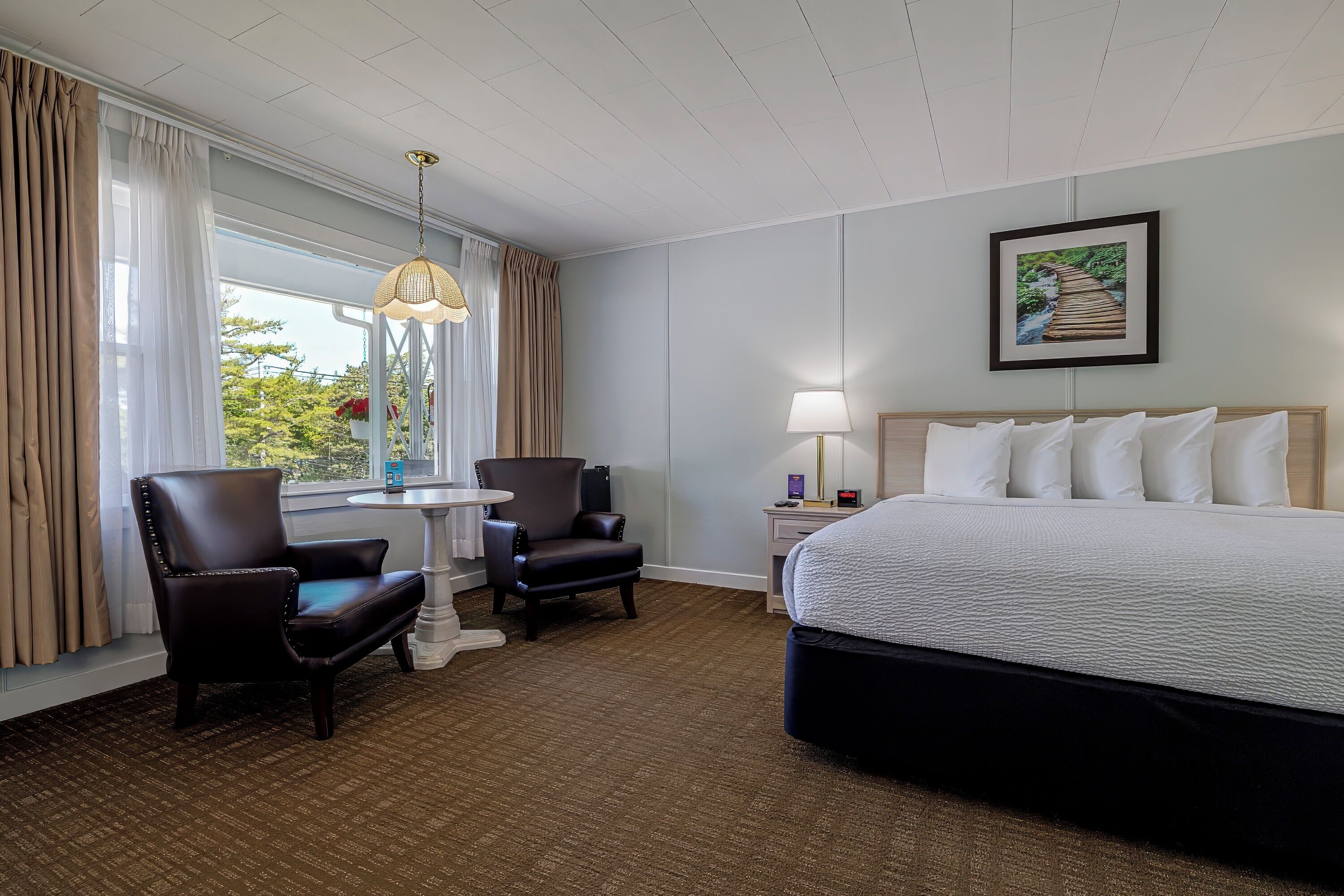 Une chambre d’hôtel avec un grand lit, deux fauteuils, une petite table, une lampe et une fenêtre avec des rideaux.