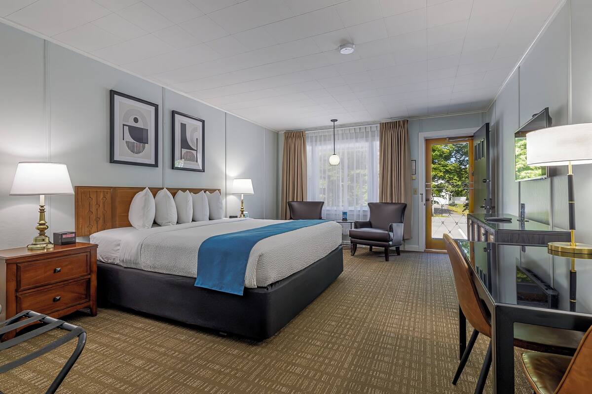 A hotel room with a large bed, two bedside tables with lamps, a desk with a chair, and a view of the outdoors through a door.
