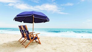 Una silla de playa bajo una sombrilla azul en una playa arenosa, con el océano al fondo.