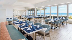 Un comedor amplio con múltiples mesas y sillas, ventanas grandes que ofrecen vista al océano, y un diseño interior moderno.