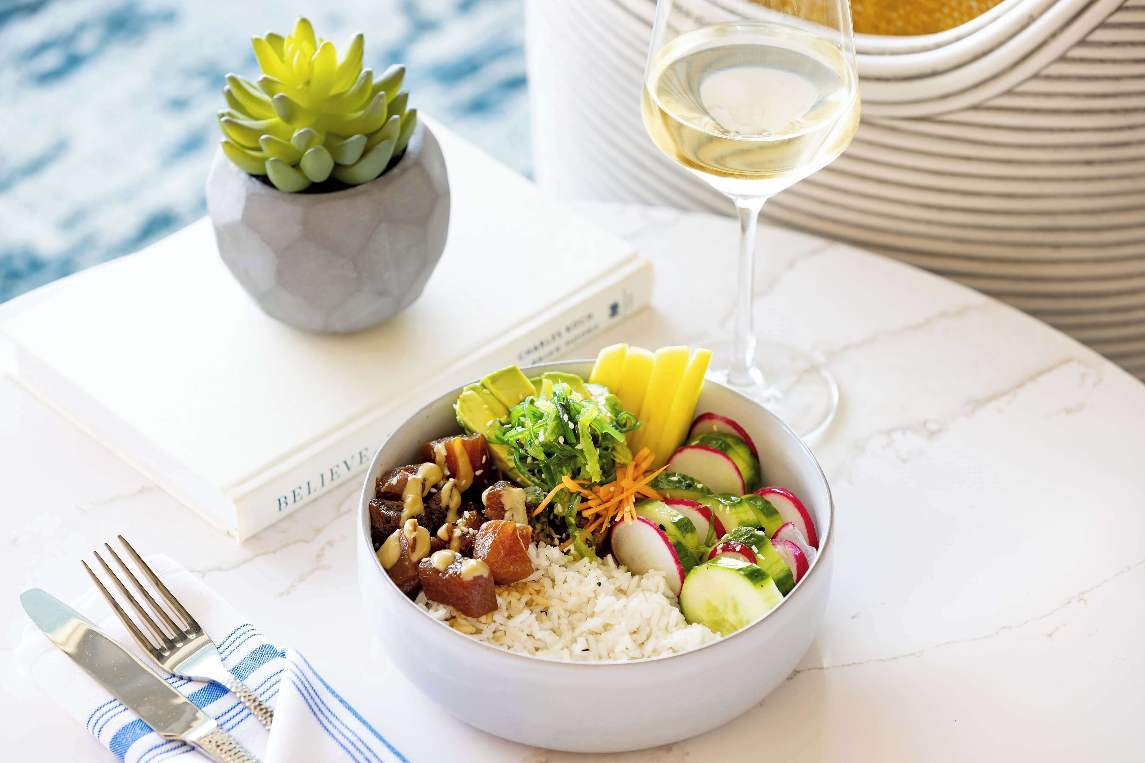 Un tazón de comida con varios ingredientes, una copa de vino blanco, un libro y una pequeña planta en maceta sobre una mesa.