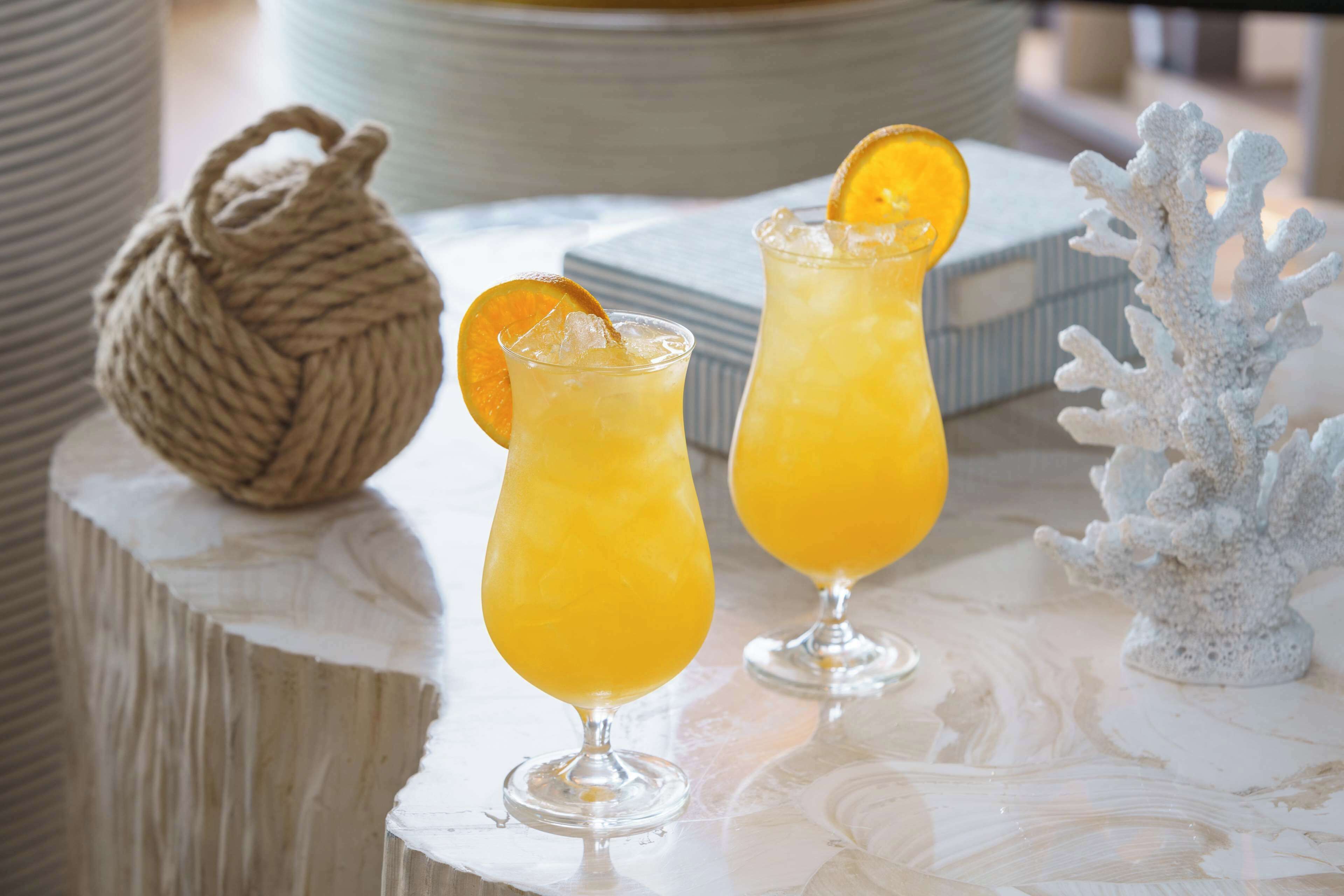 Deux verres de jus d’orange, chacun orné d’une tranche d’orange sur le bord, sont posés sur une surface en marbre. En arrière-plan, on aperçoit une boule décorative en forme de corde et une sculpture corallienne blanche.