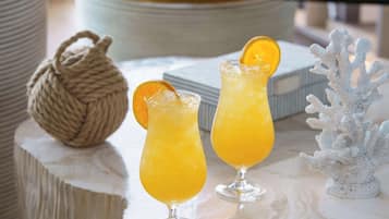 Two glasses of orange juice with a slice of orange on the rim, placed on a marble surface with a decorative rope ball and a white coral sculpture in the background.
