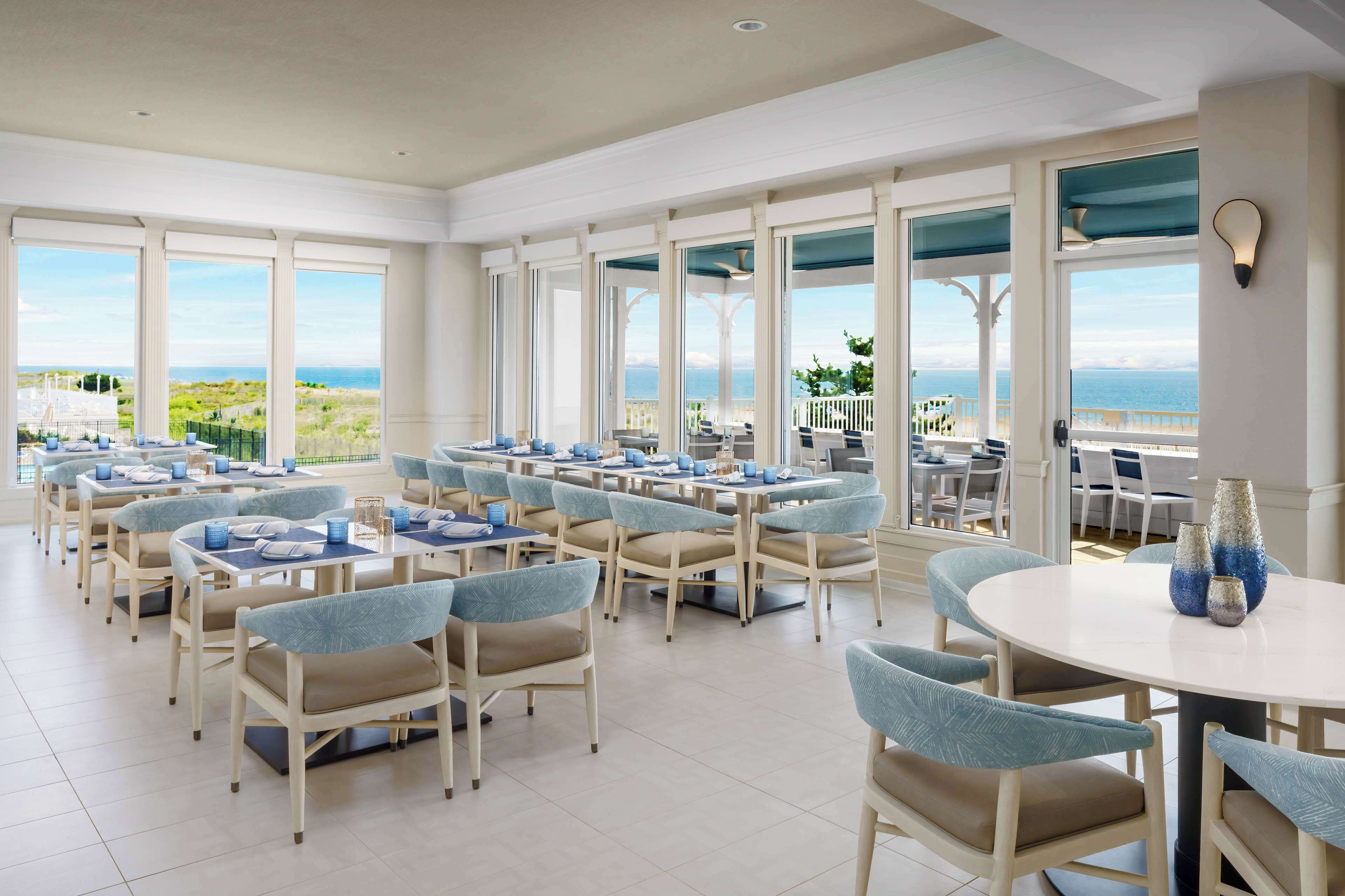 Un vaste espace dédié à la salle à manger, avec de nombreuses tables et chaises, de grandes fenêtres offrant une vue sur l’océan et un aménagement intérieur moderne.