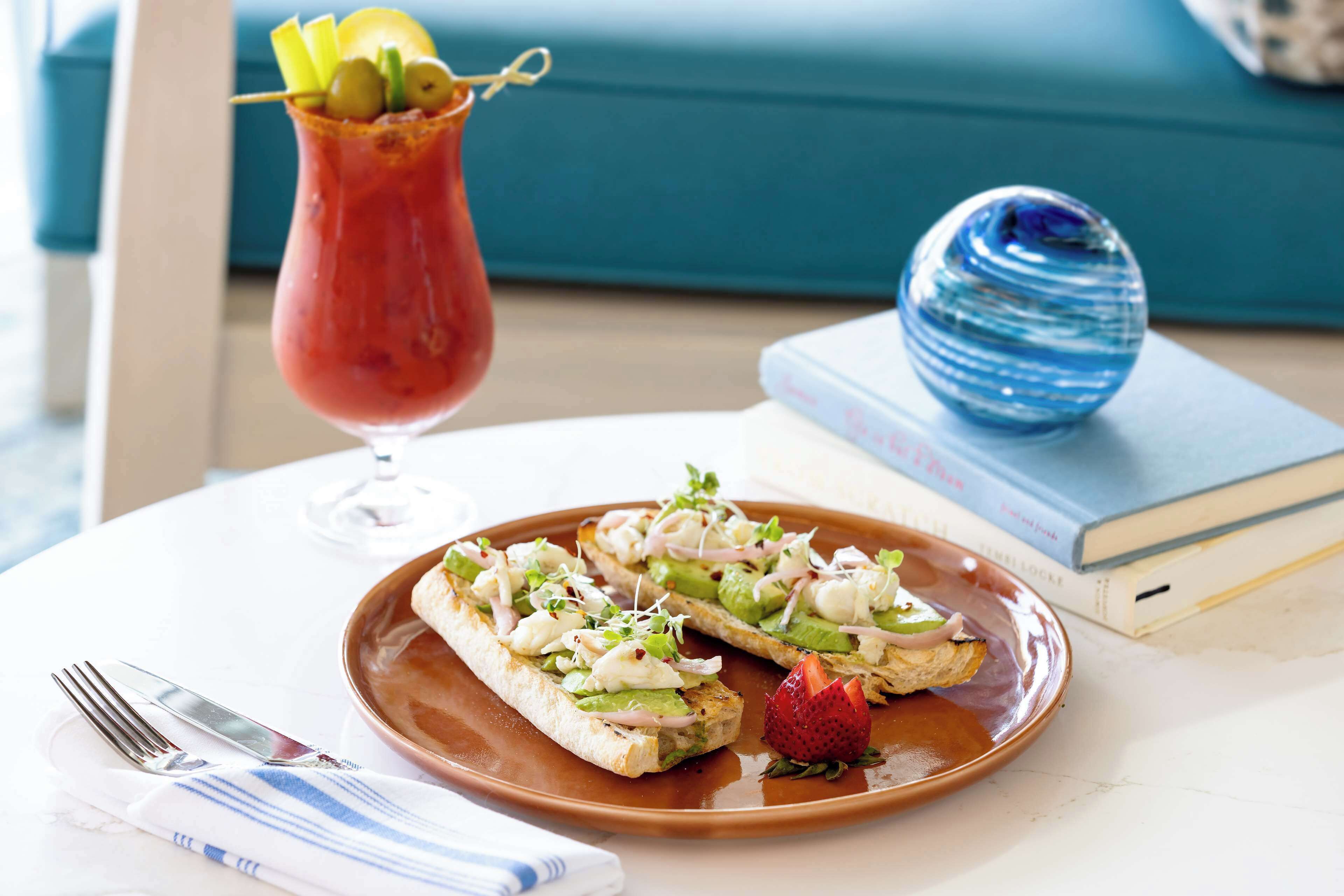 A plate with two open-faced sandwiches garnished with greens, onions, and a strawberry, a glass of red drink with a lemon slice, and a blue decorative object on a table with a book and a fork in the background.