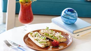 A plate with two open-faced sandwiches garnished with greens, onions, and a strawberry, a glass of red drink with a lemon slice, and a blue decorative object on a table with a book and a fork in the background.