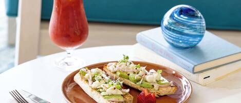 A plate with two open-faced sandwiches garnished with greens, onions, and a strawberry, a glass of red drink with a lemon slice, and a blue decorative object on a table with a book and a fork in the background.