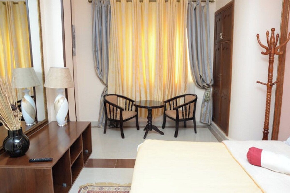 A hotel room with a bed, a small table with two chairs, a mirror, a wooden coat rack, and curtains.