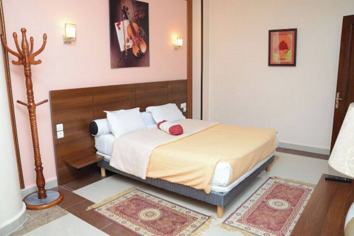A bedroom with a bed, a wooden floor lamp, a wall-mounted light, a framed picture, and a rug.