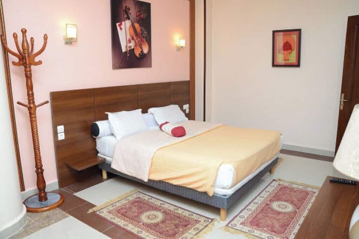 A bedroom with a bed, a wooden floor lamp, a wall-mounted light, a framed picture, and a rug.