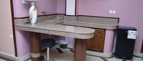 Una pequeña cocina con encimera de granito, una lámpara blanca, un refrigerador negro y una silla de madera.