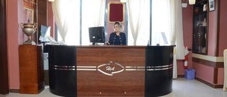 A reception desk with a wooden finish and a logo, a chandelier, and a red-framed mirror above it.