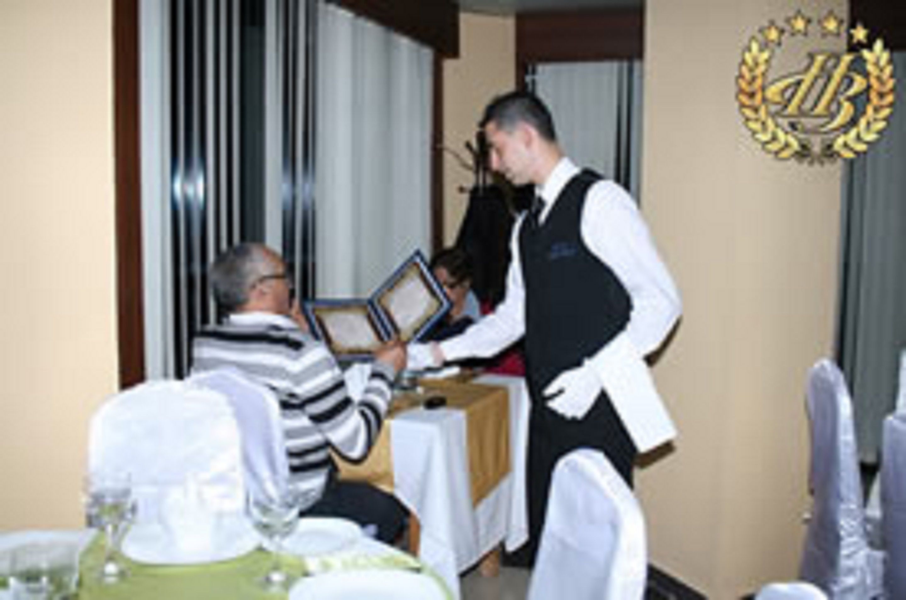 Un hombre con chaleco negro y camisa blanca entregando un diploma enmarcado a otro hombre con suéter a rayas.